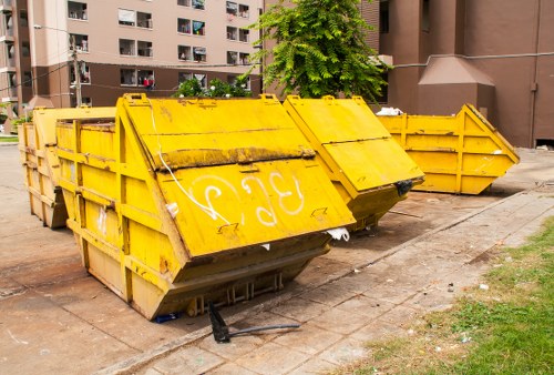 A large yellow industrial skip bin made of metal, positioned on a concrete pavement near a multi-storey residential building with dark brown exterior walls and several windows. The skip's side panels are reinforced with horizontal and vertical metal bars, and the front top is partially open with visible rust and dirt on its surface. The skip is filled with various waste materials, although the contents are not clearly visible. Behind the skip, there is a patch of grass and a small tree with lush green foliage. The surrounding area appears to be an urban environment with ample space for rubbish collection, reflective of a typical premises for rubbish removal services such as those offered by Golders Green House Clearance, operating within the London boroughs close to Golders Green postcode area. The scene is illuminated by natural daylight, emphasizing the weathered texture of the skip and the clean pavement edges. Overall, the image depicts a typical scene for waste disposal or clearance in a residential area in North West London.
