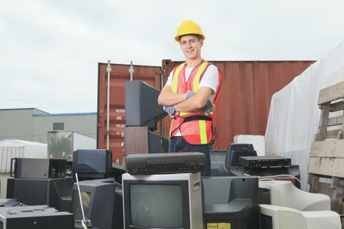 A smiling man wearing a yellow safety helmet and a high-visibility vest stands outdoors among a variety of discarded electronic equipment, including old computer monitors, printers, and other office electronics, arranged in a cluttered area. The background features a large shipping container and industrial buildings, suggesting an environment for waste collection or clearance near Golders Green. The scene captures the process of clearance, with the electronic waste spread across the ground, highlighting the type of rubbish handled by Golders Green House Clearance. Natural lighting illuminates the scene, emphasizing the textures of the electronic devices and the casual, professional appearance of the worker involved in rubbish removal services focused on electronic waste collection within the local London area.