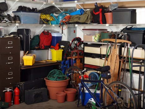 Image of a cluttered garage storage area filled with various household and outdoor items. In the foreground, there is a blue bicycle leaning against a shelving unit, with a black and blue portable speaker nearby. Several empty terracotta plant pots and a green garden hose are stacked on a yellow tray on a black workbench, which also holds a red toolbox and a small white plastic container. Behind the workbench, a white shelving unit is filled with plastic storage boxes, some transparent and others opaque, containing various miscellaneous objects. On the top shelf, there are trousers, bags, and some black footwear. To the right of the bicycle, a wooden garden chair is partially visible, leaning against the shelving unit. The overall scene is illuminated by indoor lighting, emphasizing the mixture of household clutter and outdoor tools, typical of waste or rubbish that a rubbish removal service like Golders Green House Clearance might assist with in the local area near London.