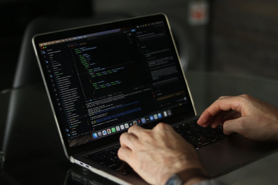 A person seated at a desk is using a laptop computer displaying multicolored lines of code on the screen, with their right hand holding a pen or stylus over the device's touchpad. The individual is pa